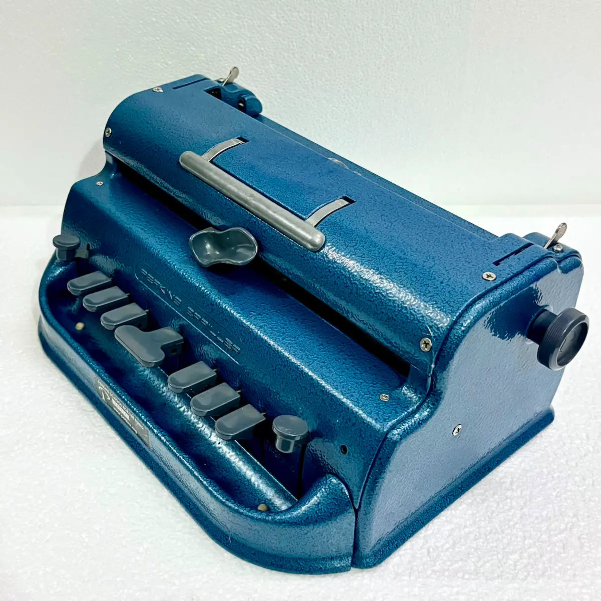 Three-quarter view of a blue Perkins Braille typing machine showing carriage, keys and textured metal body, RR Typewriter Mart