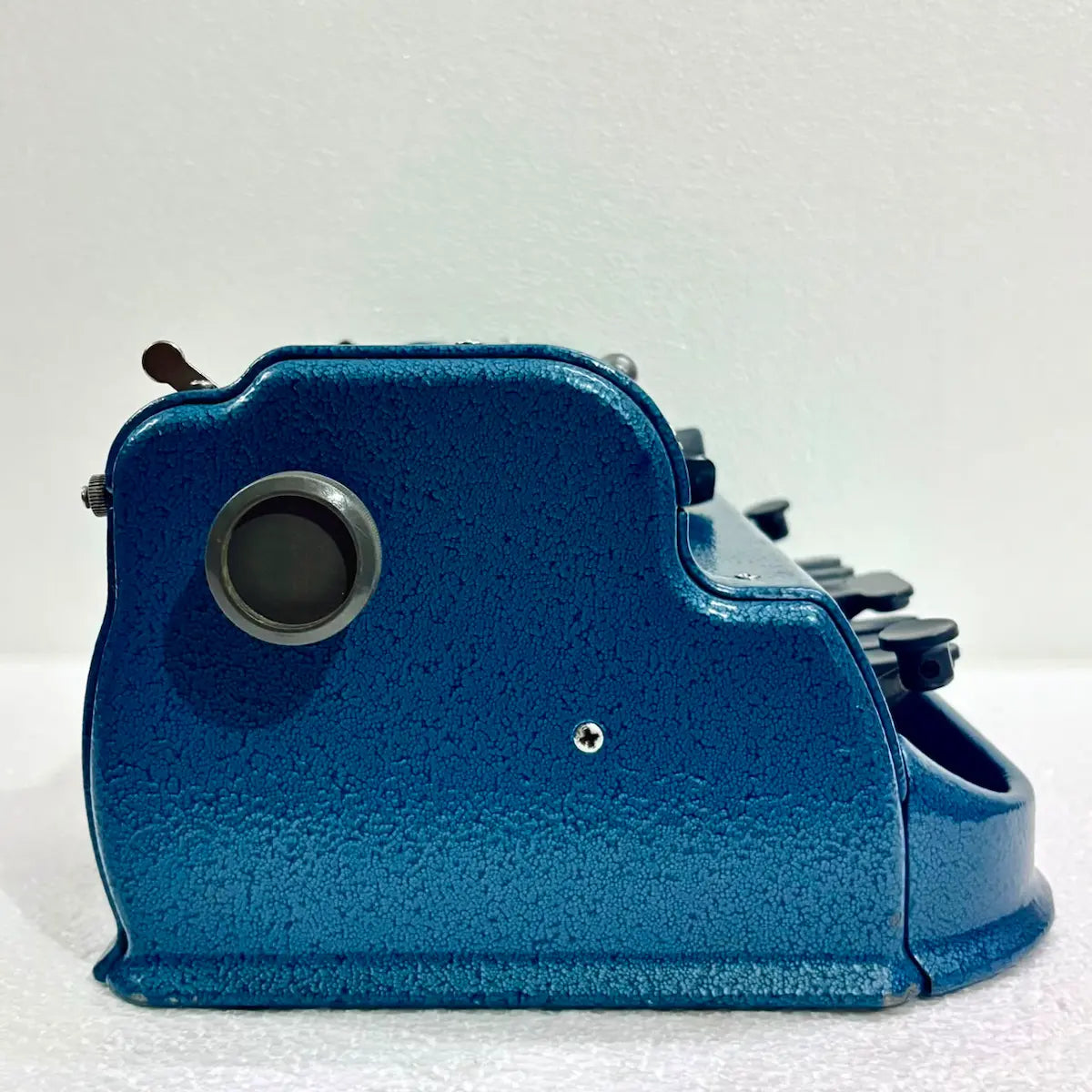 Side view of a blue Perkins Braille Typewriter showing its textured metal body and carriage knob, from RR Typewriter Mart