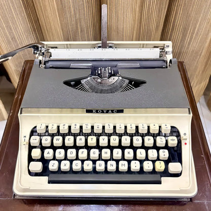Kovac M-101A Japanese portable typewriter in original grey and ivory white, with cream keys and exposed typebars, photographed on a wooden table | RR Typewriter Mart