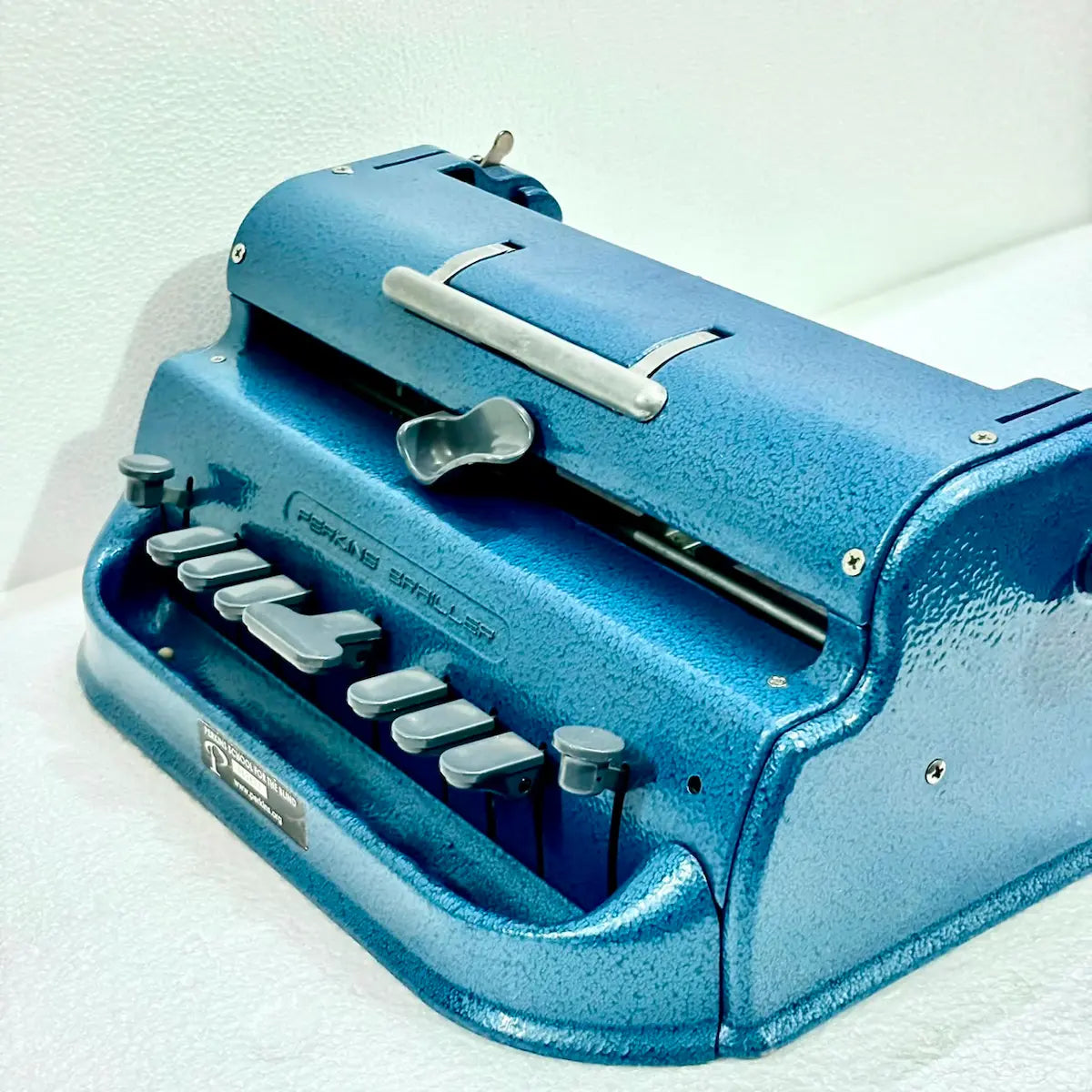 Three-quarter view of a blue Perkins Brailler typewriter with six-key layout and paper guide, an original Braille typing machine at RR Typewriter Mart
