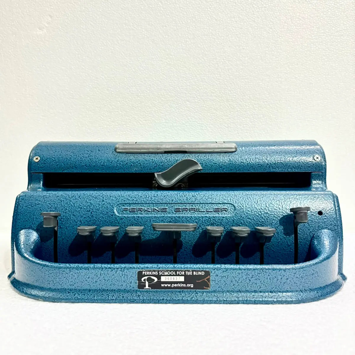 Front view of a blue Perkins Braille Typewriter showing six Braille keys and textured metal body, photographed by RR Typewriter Mart