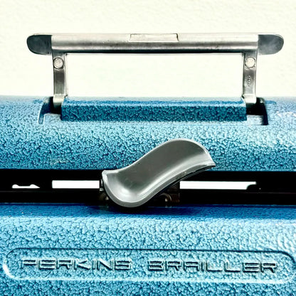 Close-up of a blue Perkins Brailler typewriter showing its handle and embossed logo, an imported Perkins Braille machine at RR Typewriter Mart