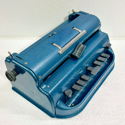 Three-quarter view of a blue Perkins Braille Typewriter showing keys, carriage and textured body, an imported Braille typing machine at RR Typewriter Mart